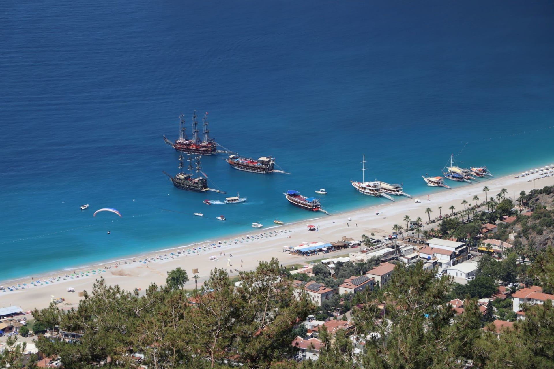 Ölüdeniz Blue Lagoon framed by pine-covered mountains, a paraglider descending in the distance against a cloudless July sky