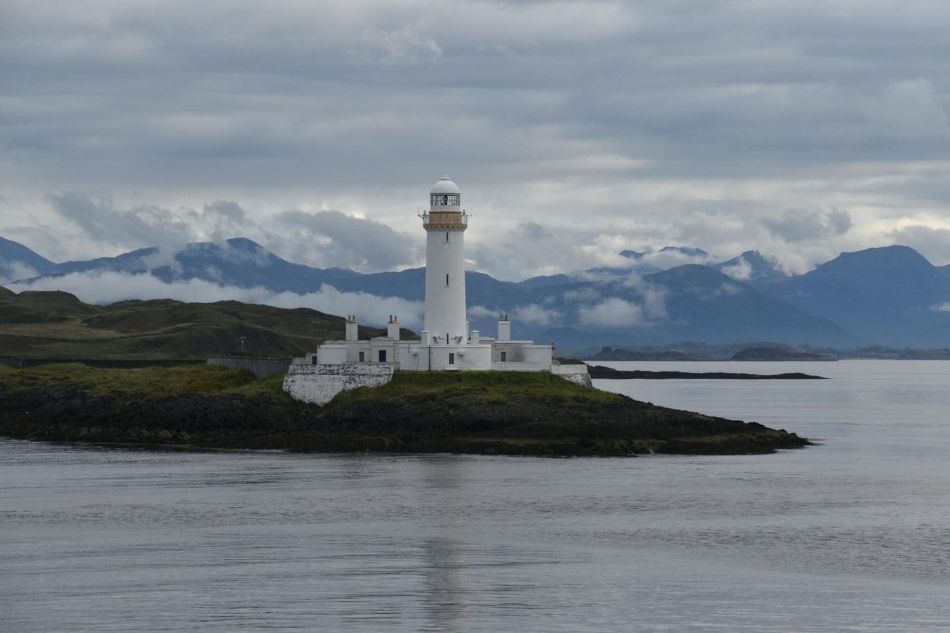 The horseshoe harbour of Oban at dusk, fishing boats moored along the stone pier with the colonnaded McCaig's Tower rising on the hill behind, Kerrera island dark against a pale Atlantic sky.