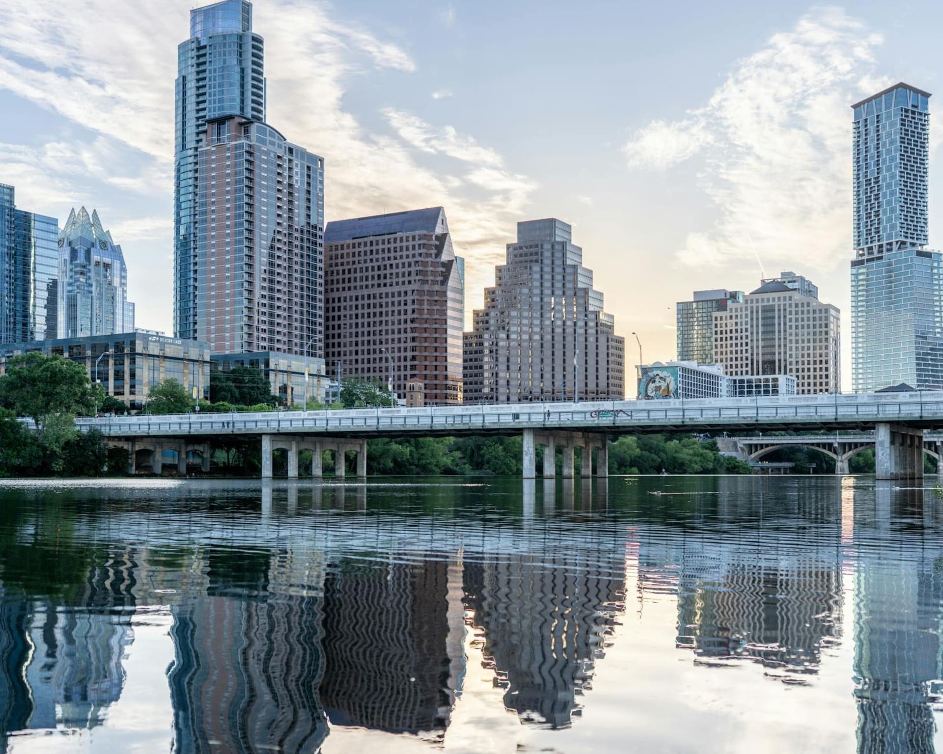 Austin's downtown skyline glowing amber at dusk, the Congress Avenue bridge arching over Lady Bird Lake with bats beginning to rise in dark ribbons against the fading sky.