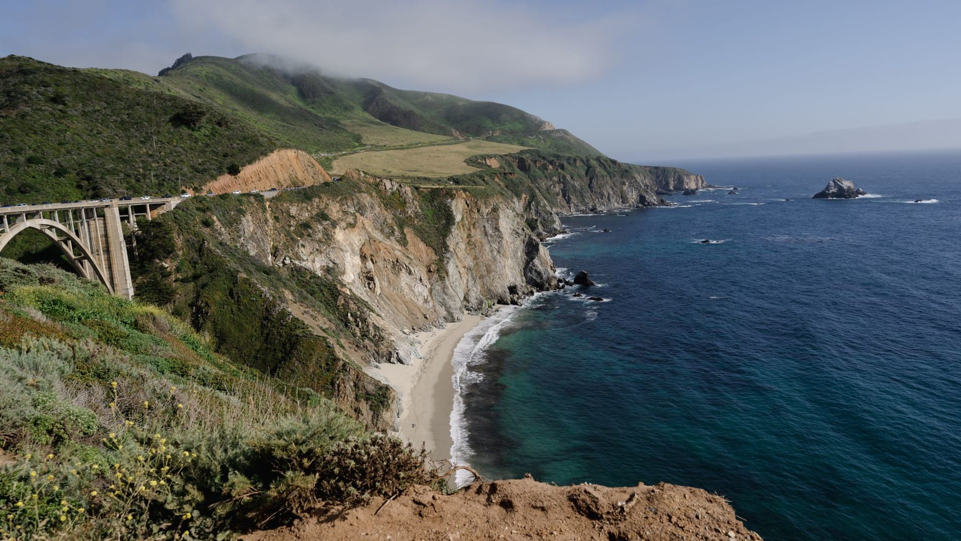A sweeping view of Big Sur's rocky coastline from Highway 1, with golden cliffs dropping into a churning teal Pacific and wisps of coastal fog clinging to the Santa Lucia range above.