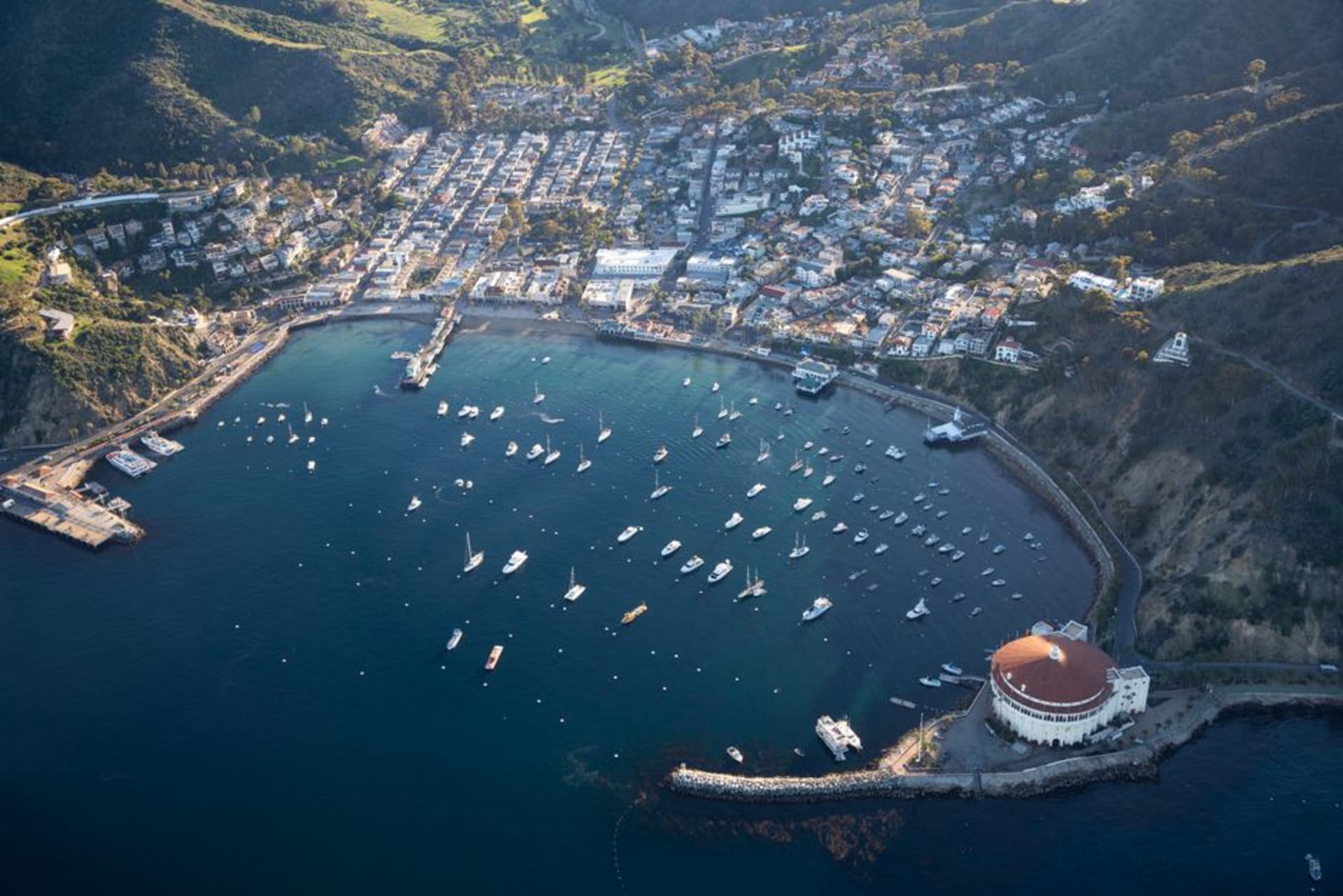 The harbor town of Avalon on Catalina Island with pastel buildings, moored boats, and the round Casino building against green hills