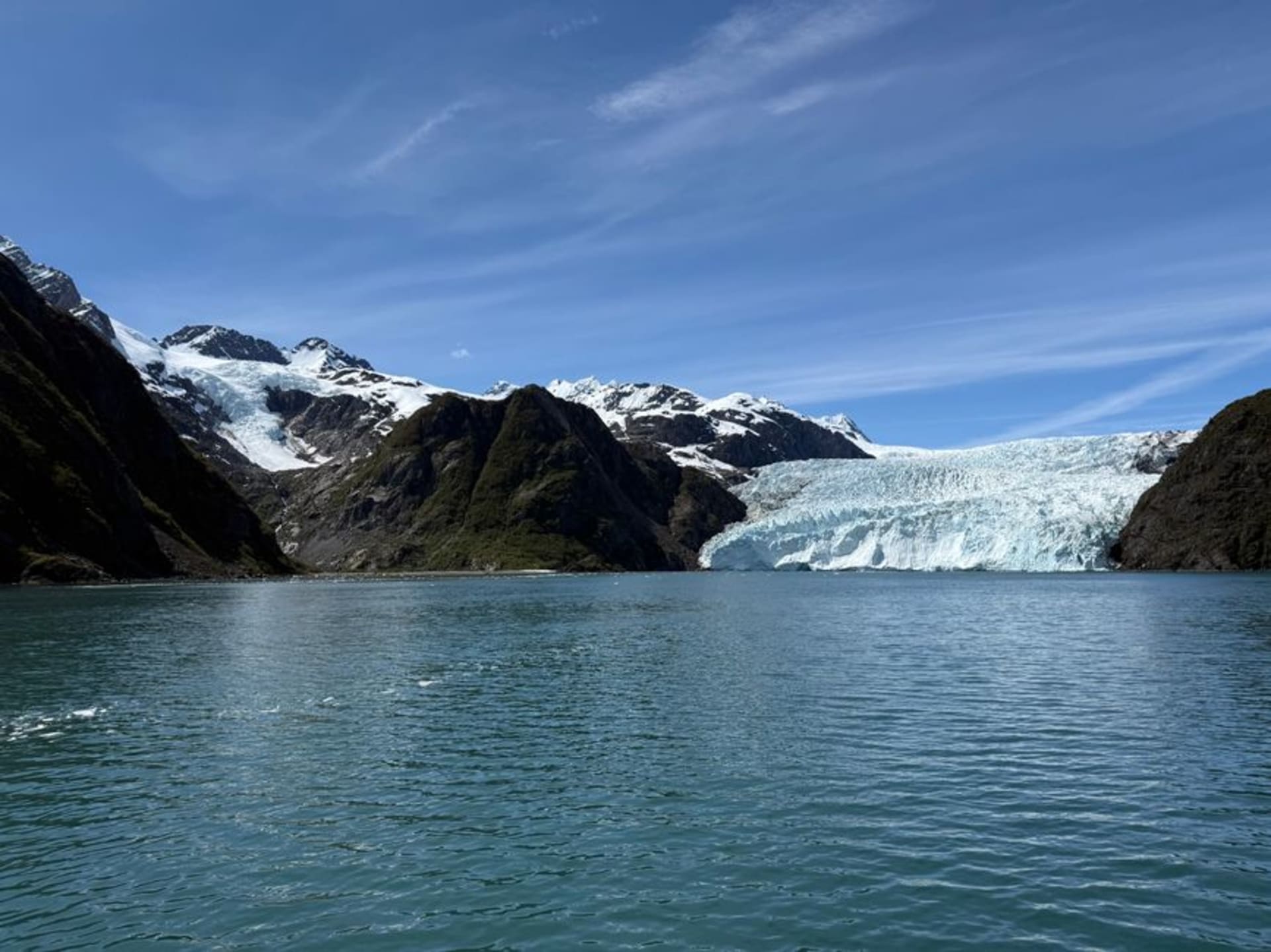A tidewater glacier calving into the green water of a fjord under mountains in Kenai Fjords