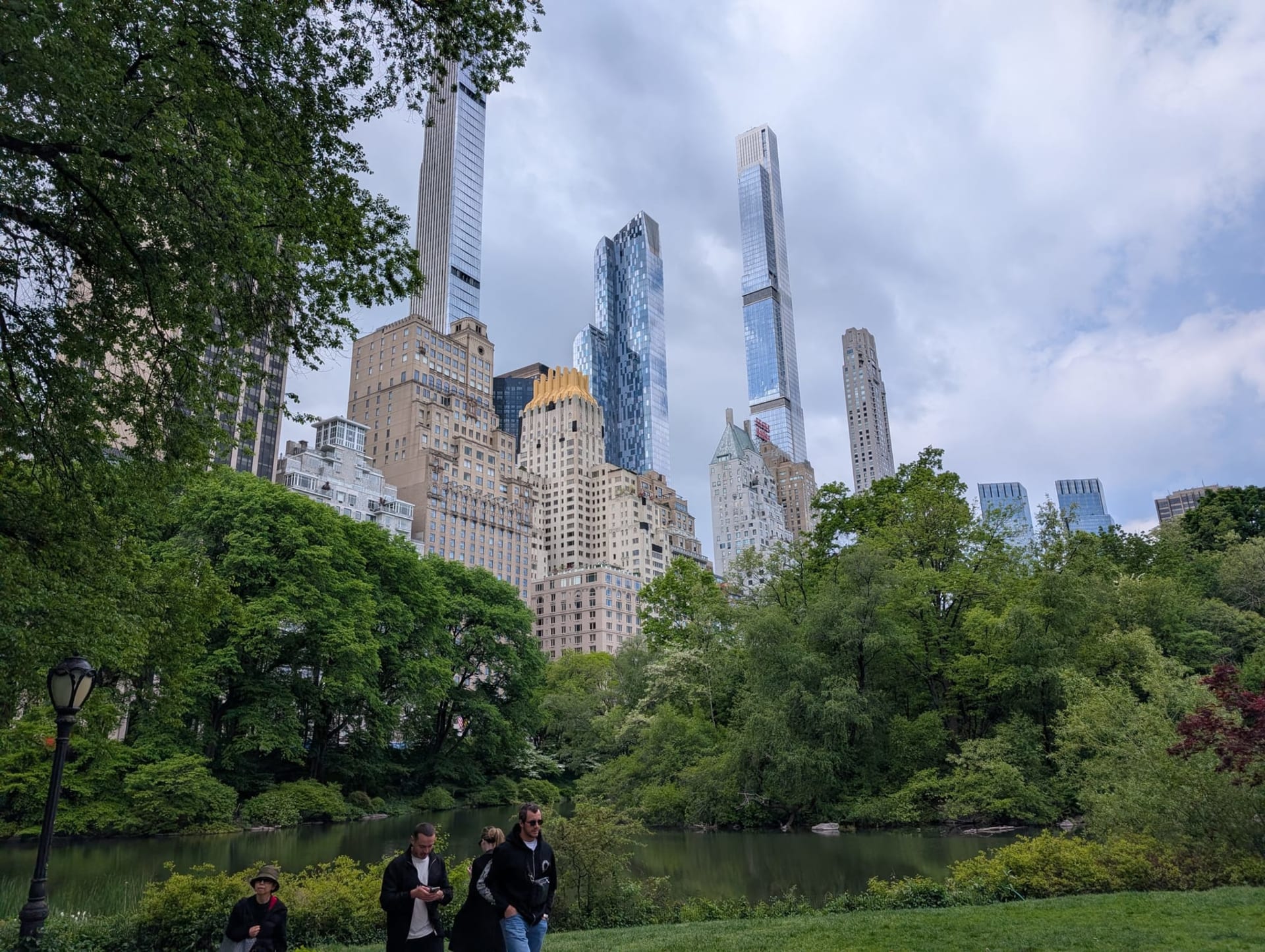 Central Park's Pond in early May — green trees framing a still stretch of water, the midtown towers rising beyond the treeline: One57, 432 Park, and the needle-thin supertalls floating above the leaves