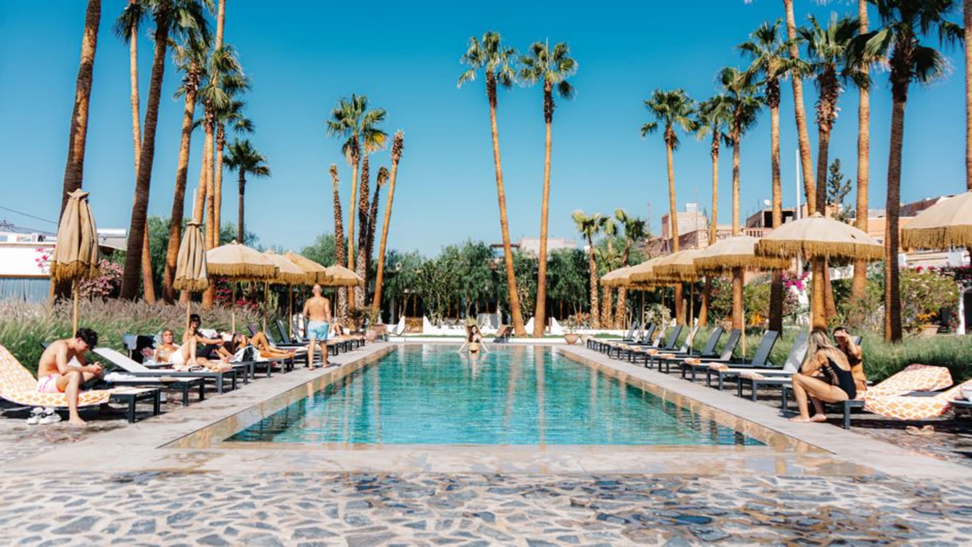 A midcentury modern house in Palm Springs with a turquoise pool and tall palm trees, the barren San Jacinto mountains rising steeply behind.