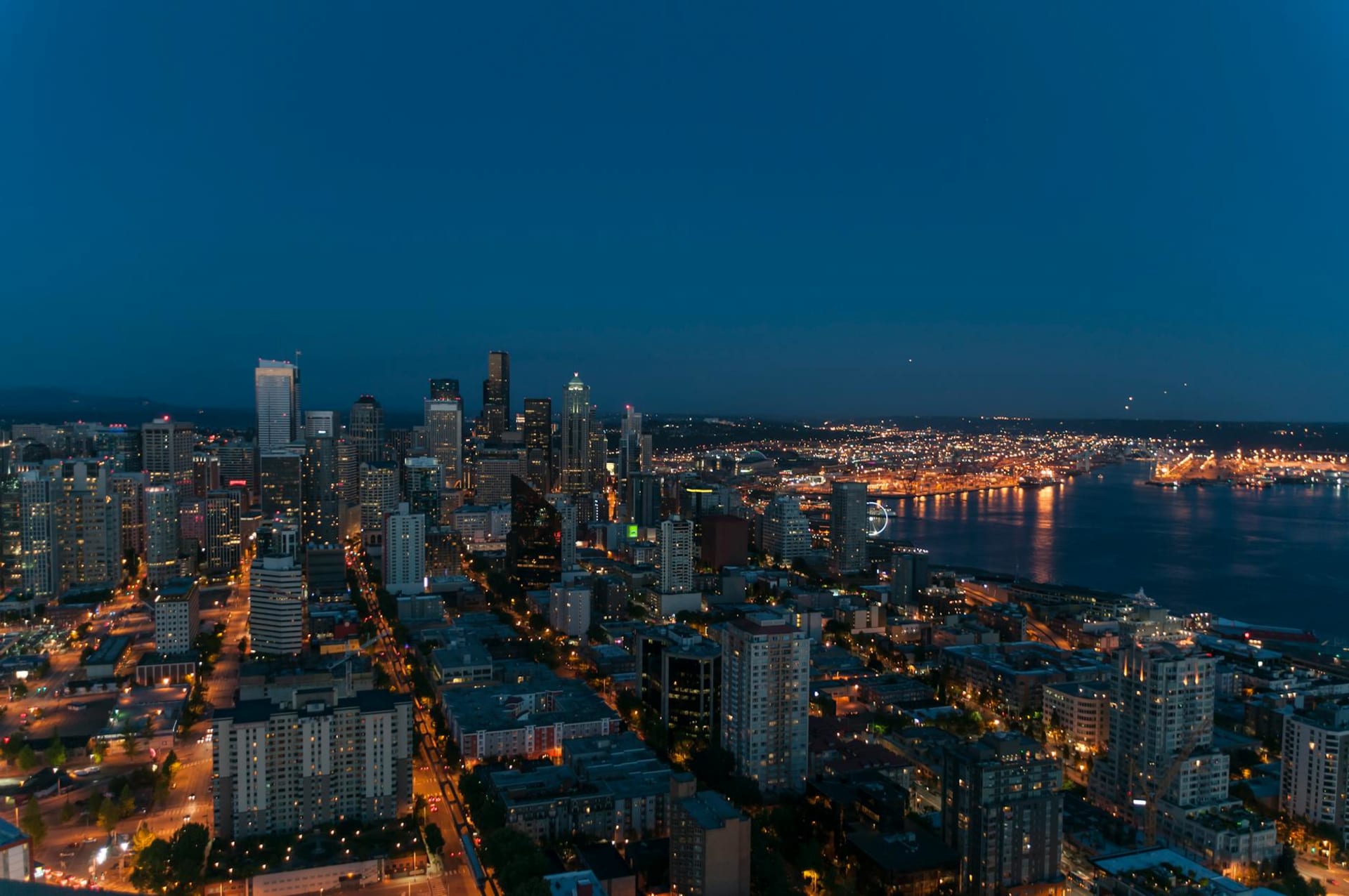 The Space Needle rising above Elliott Bay at dusk, city lights reflected on wet pavement with the Olympic Mountains fading into cloud behind it