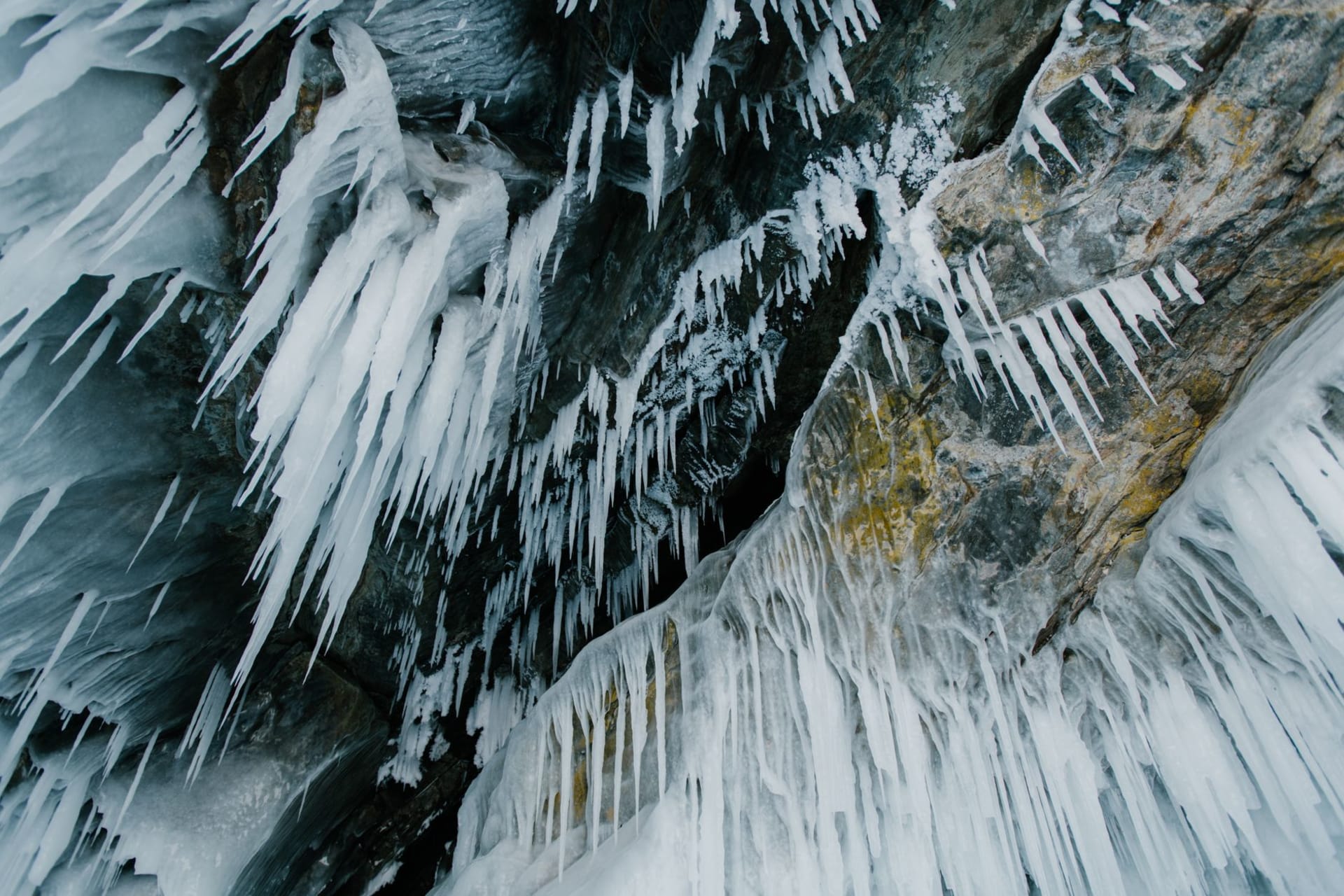 Ice stalactites illuminated in blue-green light inside a cathedral-sized cavern of the Kungur Ice Cave, their reflections pooled on a frozen floor