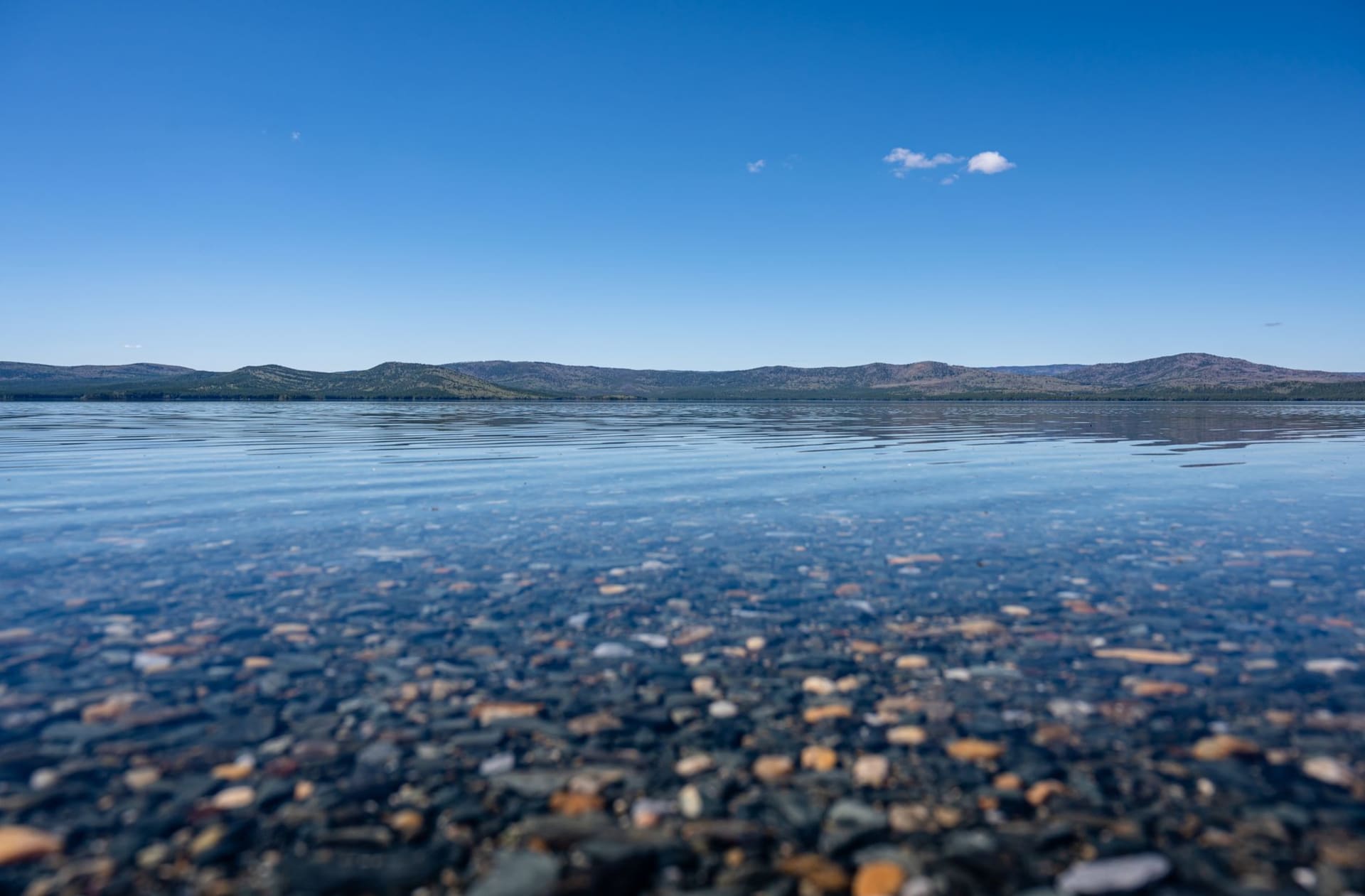 Lake Turgoyak seen from a granite boulder on the eastern shore, the water shifting from transparent turquoise in the shallows to deep cobalt, pine forest reflected perfectly in the still surface