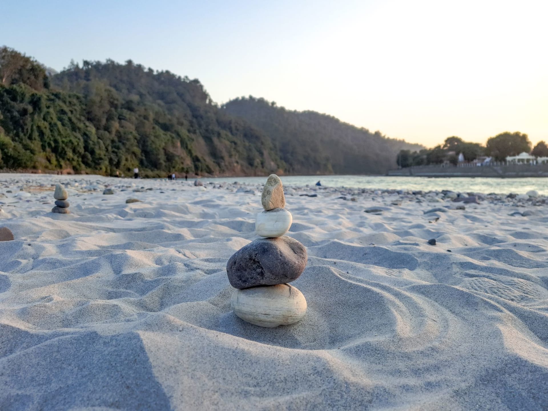 Cairns of stacked river stones on a sandy Ganges riverbank in Rishikesh, with the sacred river glinting in the background