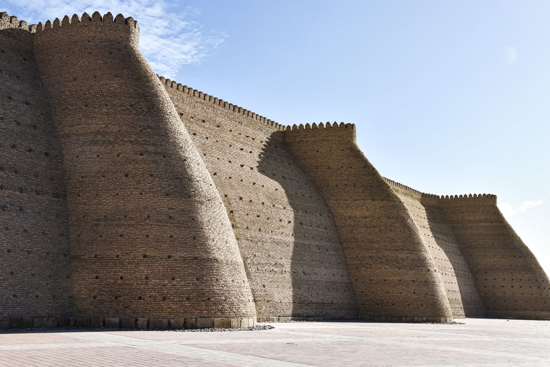 The Kalon Minaret rising above Bukhara's flat rooflines at dawn, its brickwork warm against a pale desert sky, pigeons circling the upper balcony