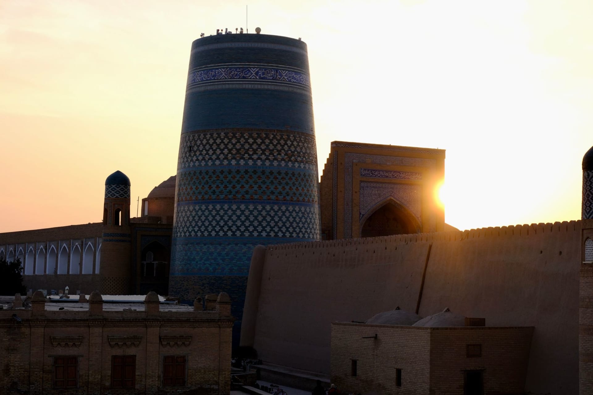 The Kalta Minor turquoise minaret stub and the Islom-Hoja minaret rising above Khiva's ancient mud-brick walls at sunset, the Kyzylkum desert extending to the horizon