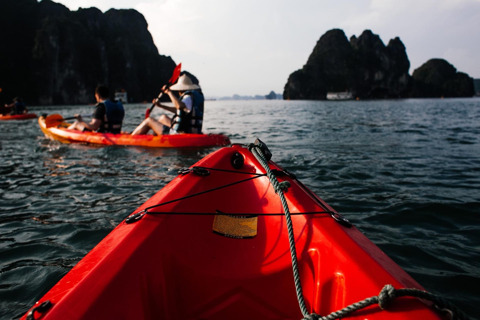 A yellow kayak gliding through a narrow limestone cave opening onto a hidden emerald lagoon, the karst walls rising sheer and moss-draped above glassy water in the early morning mist of Halong Bay.