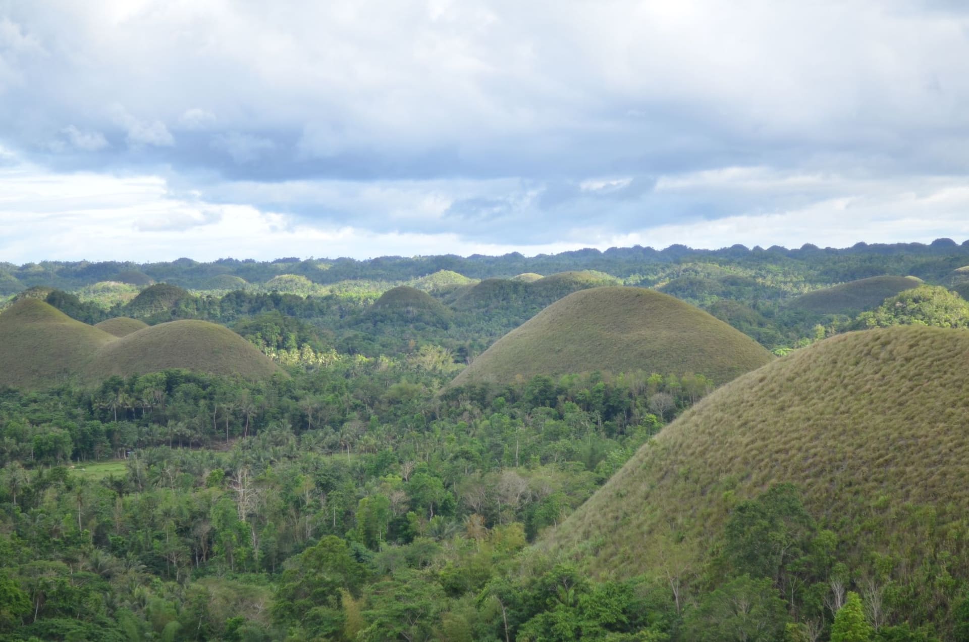 Panoramic view of the Chocolate Hills of Bohol — hundreds of perfectly rounded grass-covered mounds stretching to the horizon under a dramatic cloudy sky