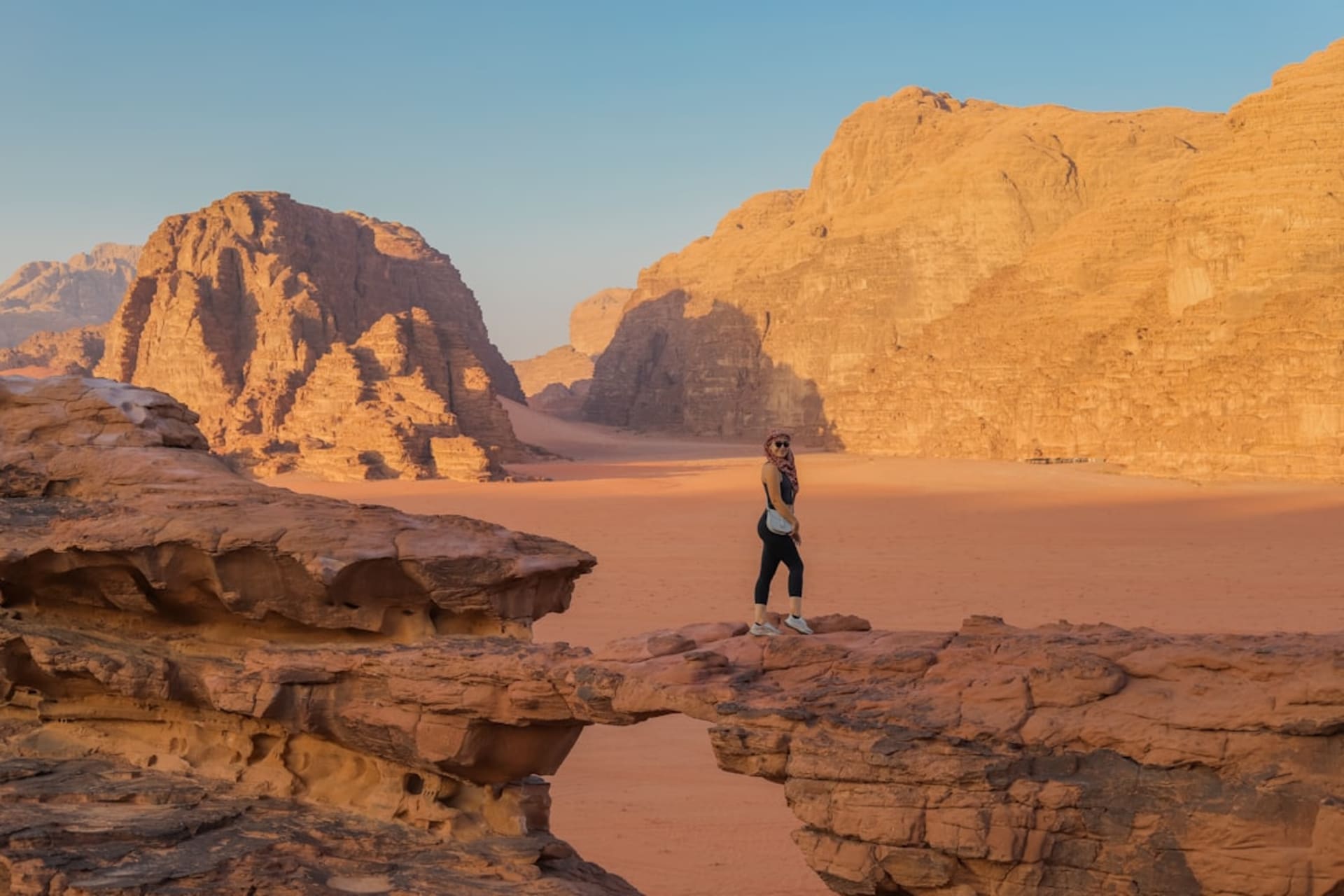 A massive sandstone arch spanning a gap high above the Wadi Rum desert floor, orange rock against a deep blue sky with the valley stretching to the horizon