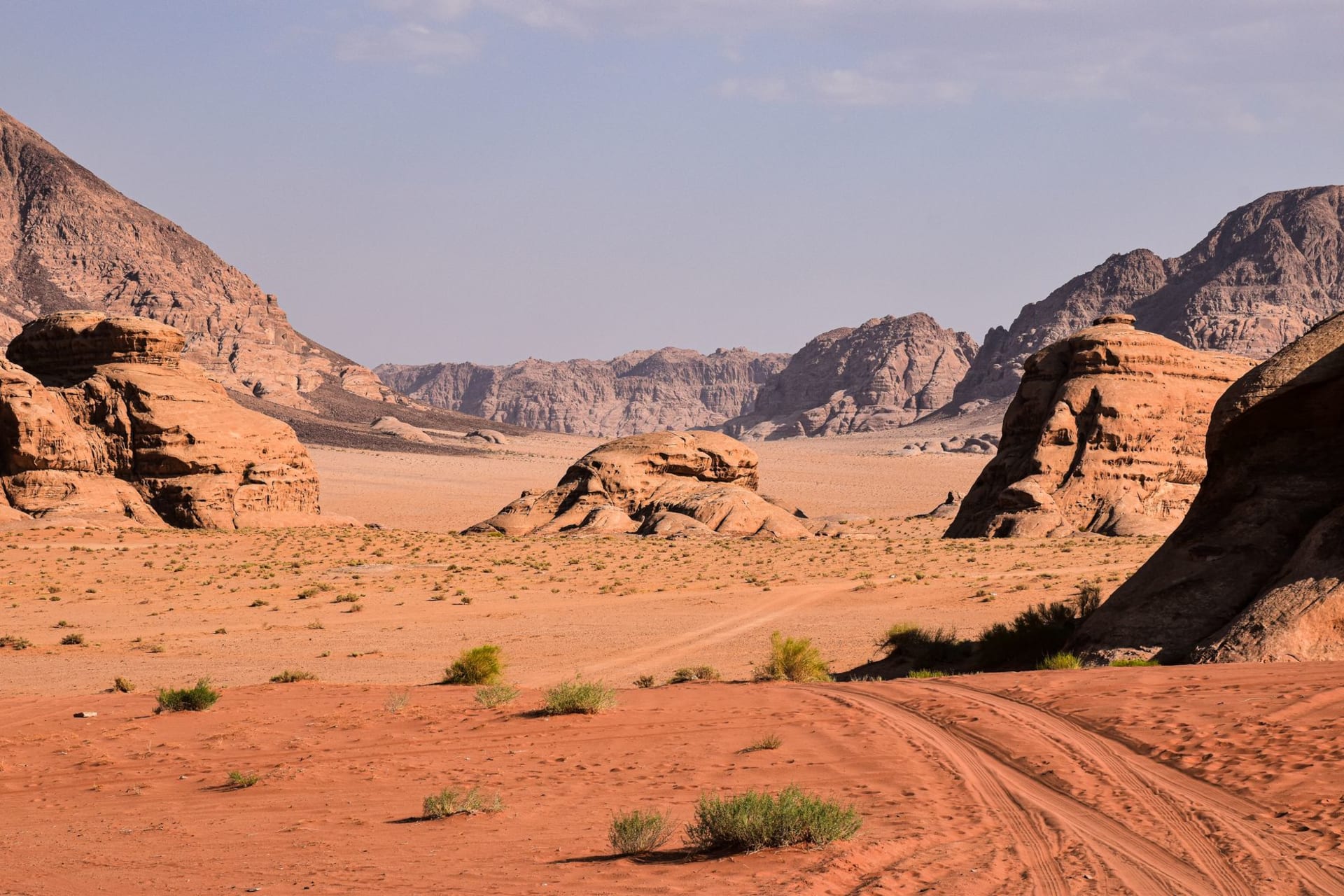 Rust-red desert floor of Wadi Rum stretching toward layered sandstone mountains, with tire tracks cutting through the sand and sparse desert scrub under a pale blue sky