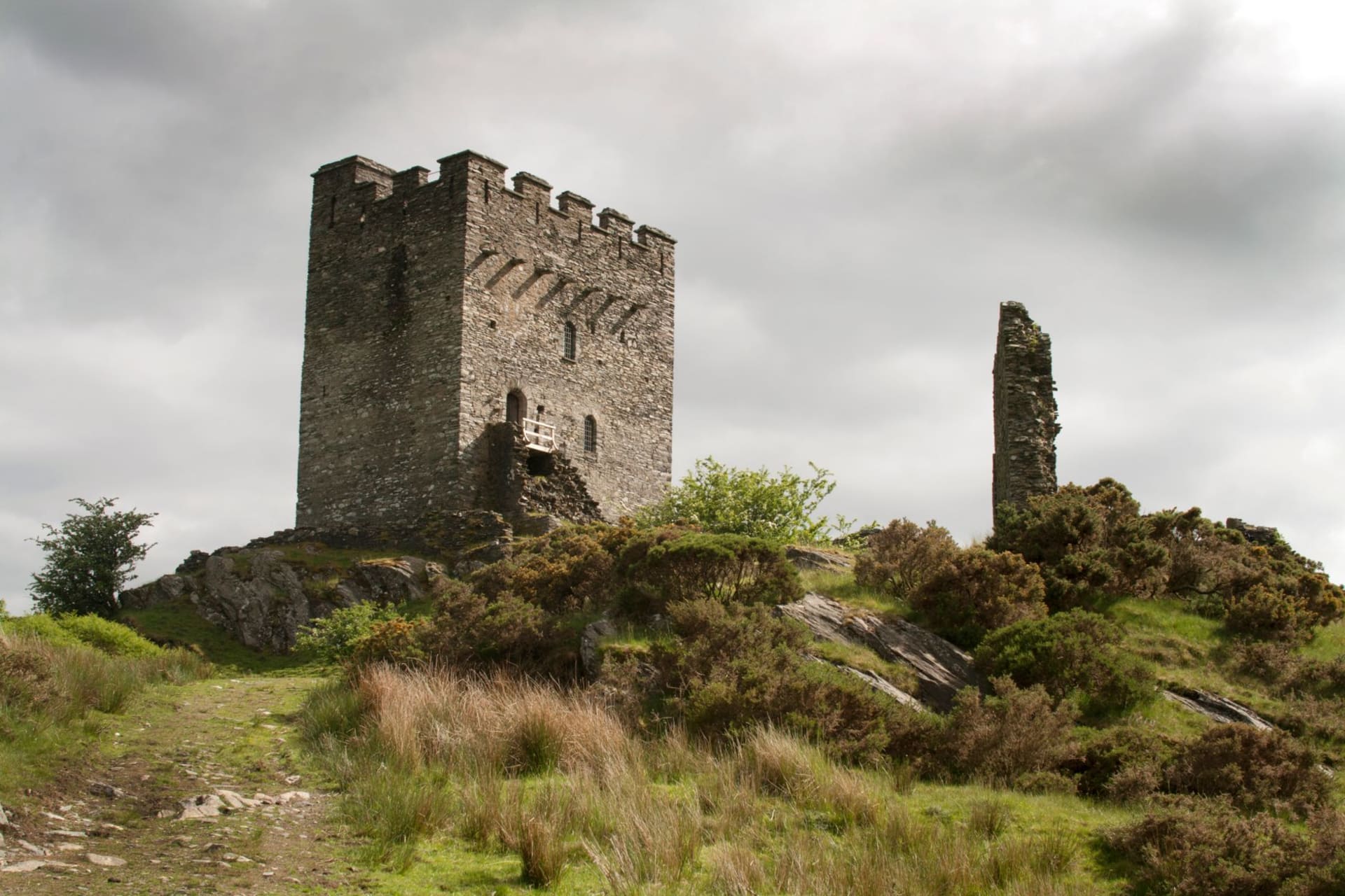 Dolwyddelan Castle, a medieval stone tower perched on a green hillside in the Conwy Valley, with dramatic Welsh mountains rising behind it under overcast skies