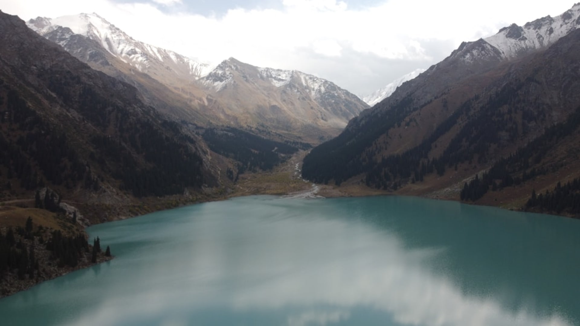 Kanas Lake glowing deep turquoise between pine-forested slopes in autumn, with golden larch trees reflected in the still water and a wooden Tuvan cabin visible on the far shore