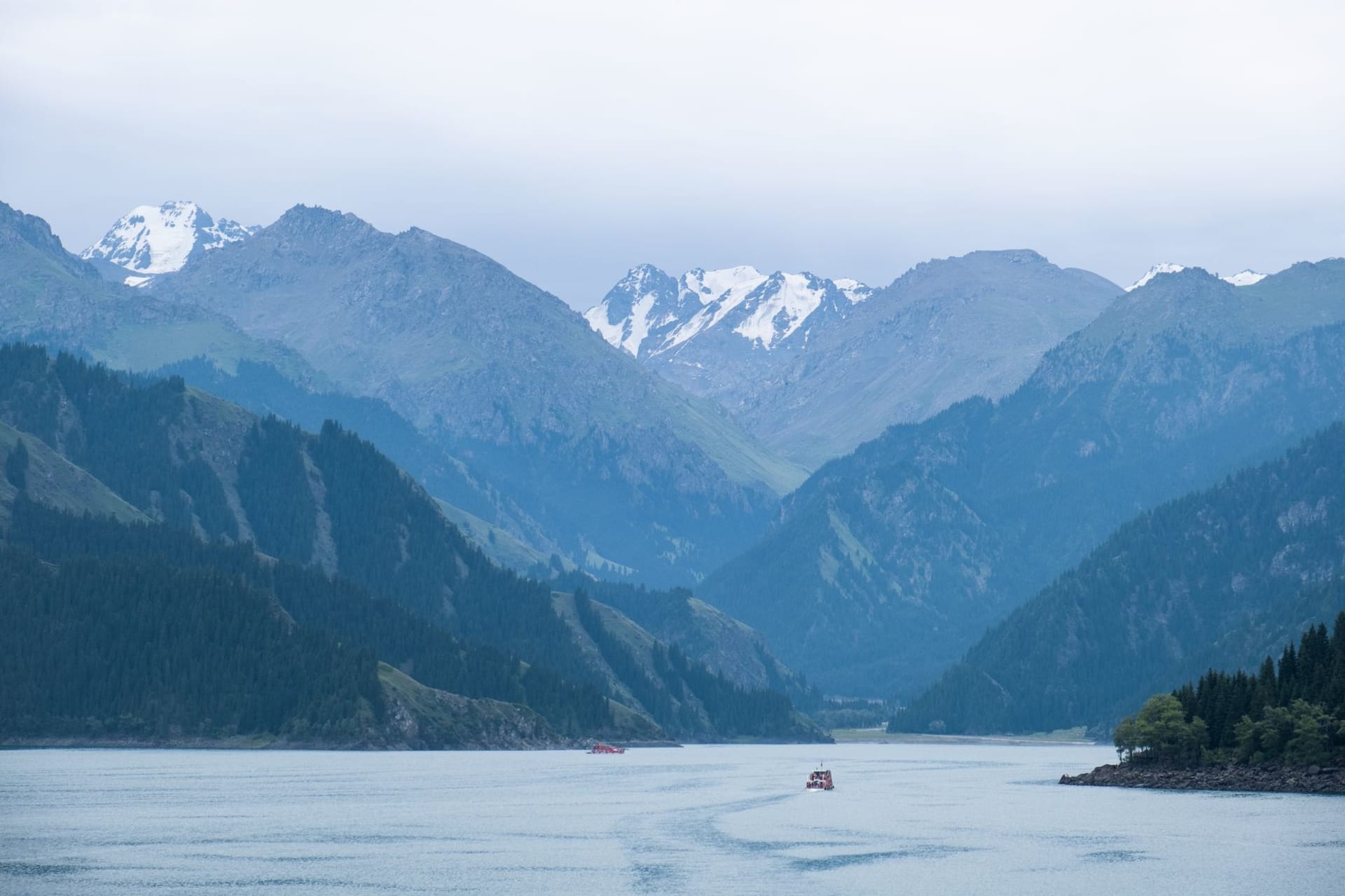 Le lac Sayram miroitant d'un bleu cobalt profond sous un ciel d'orage spectaculaire, avec des pentes couvertes de fleurs sauvages jaunes et violettes descendant vers l'eau et des sommets mouchetés de neige au-delà