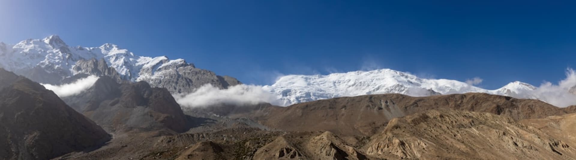 The eroded earthen towers of Tashkurgan's Stone City rising from a green wetland at dusk, with Kongur Tagh's massive snowy bulk filling the entire background sky