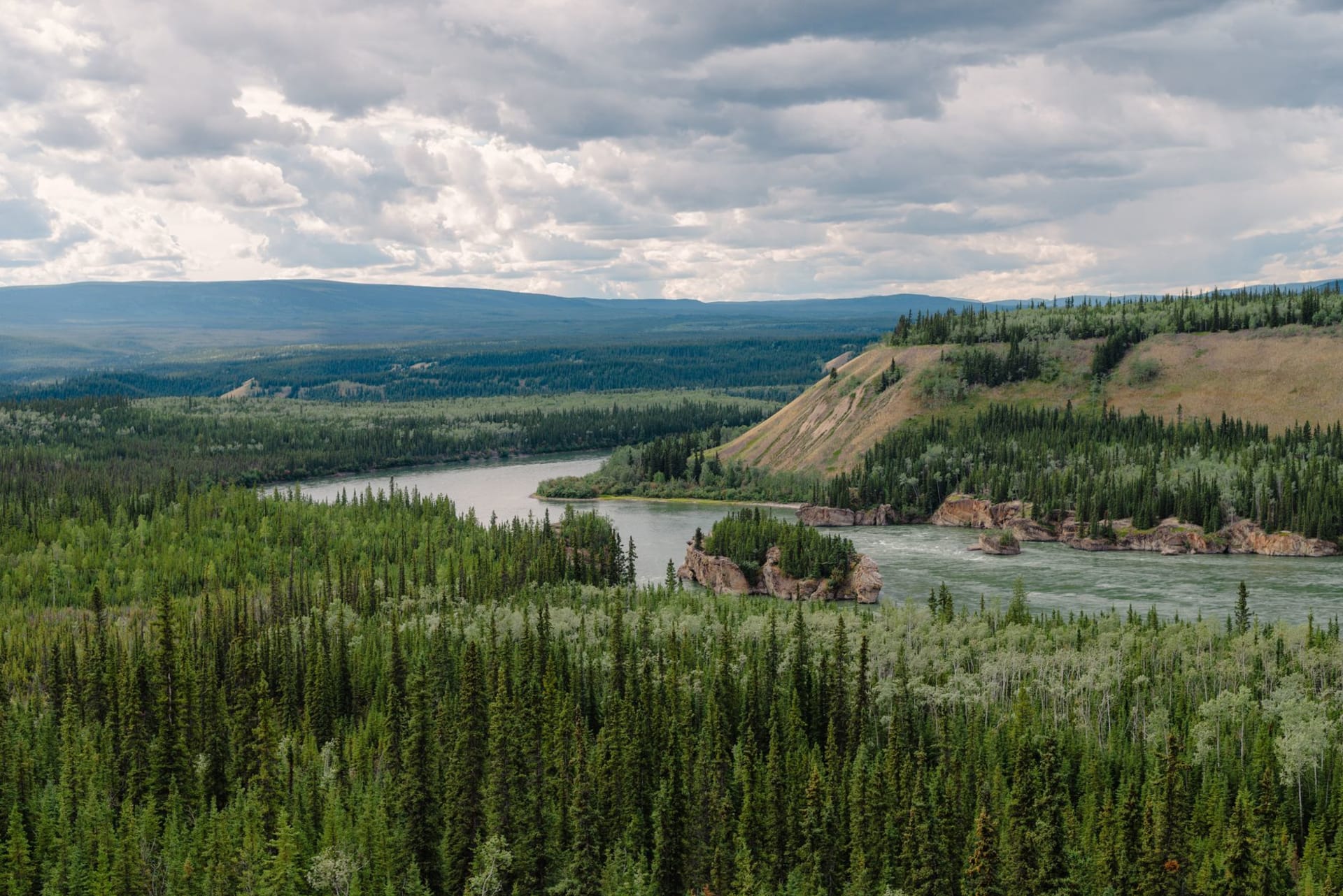 The Dempster Highway stretching north across an open tundra plateau under a dramatic cloud-streaked sky, the Richardson Mountains visible on the horizon