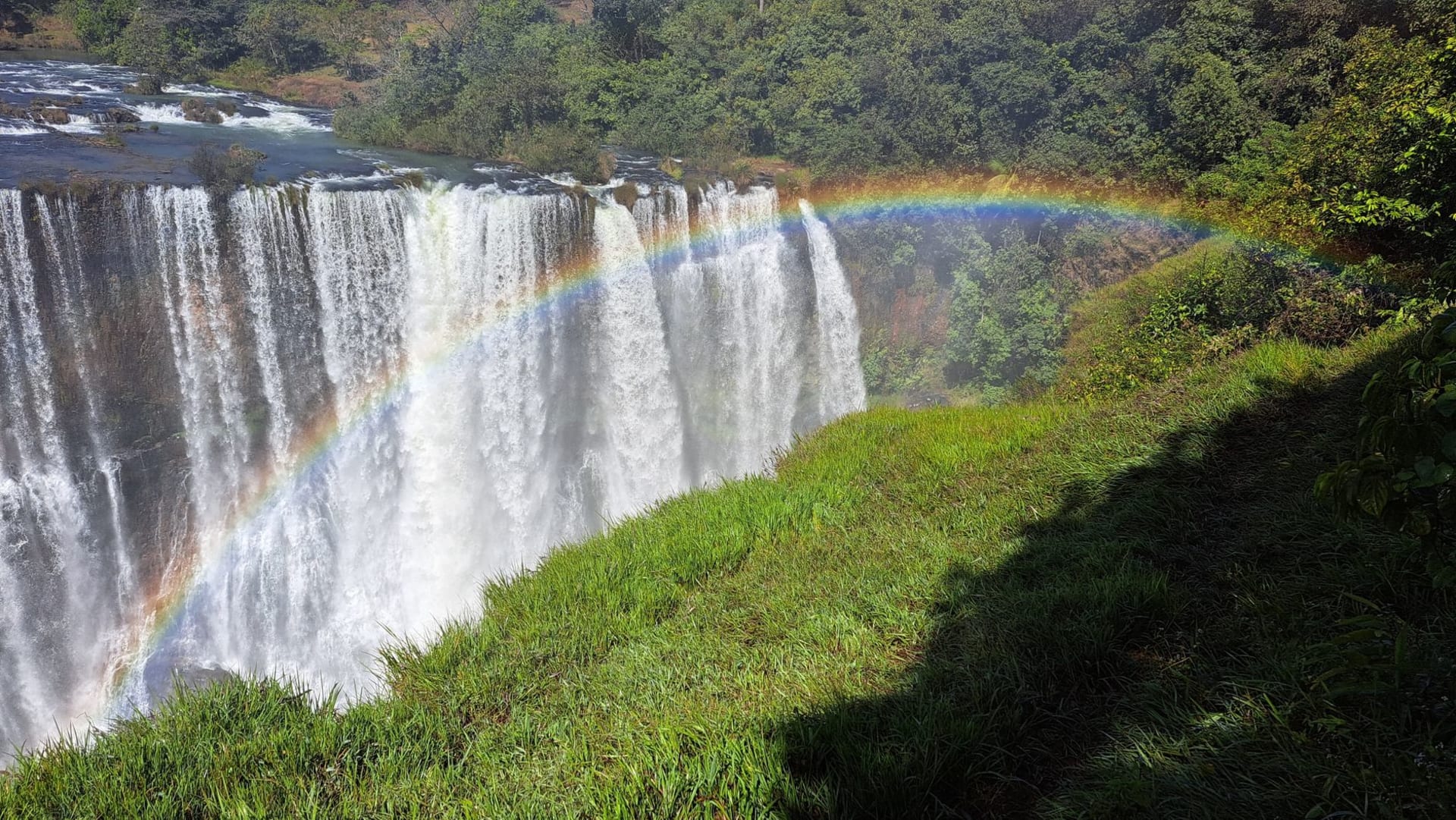 Las cataratas Victoria irrumpiendo en un muro de niebla blanca sobre el cañón del Zambeze, con un arcoíris atravesando el spray al mediodía
