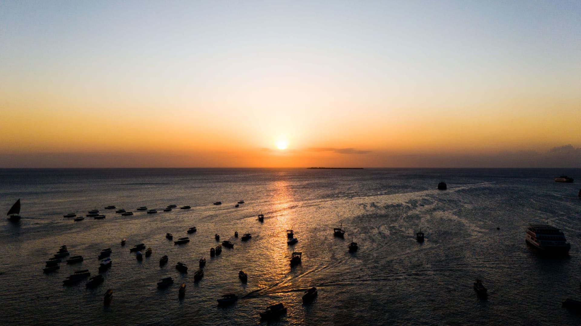 Wooden dhows anchored in calm waters at sunset off Stone Town, Zanzibar, with a warm orange sky reflected on the sea