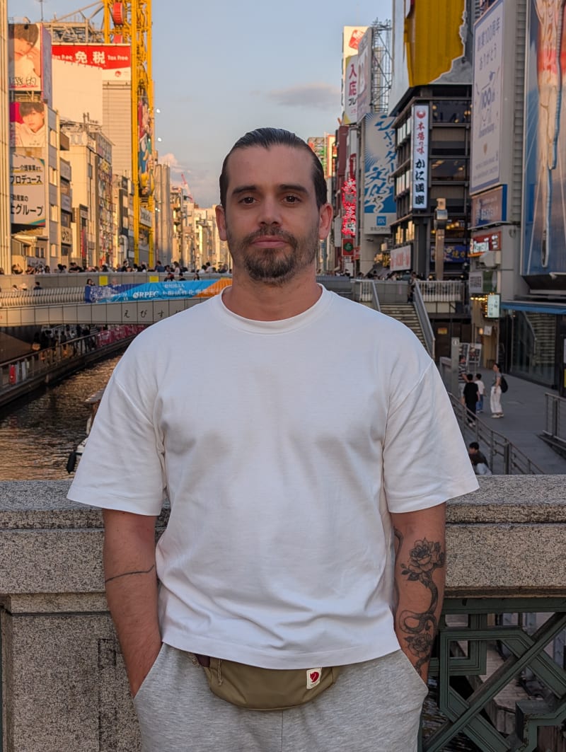 Pierre standing on a bridge over the Dotonbori canal in Osaka at dusk, the neon signs and billboards of the street lit up behind him