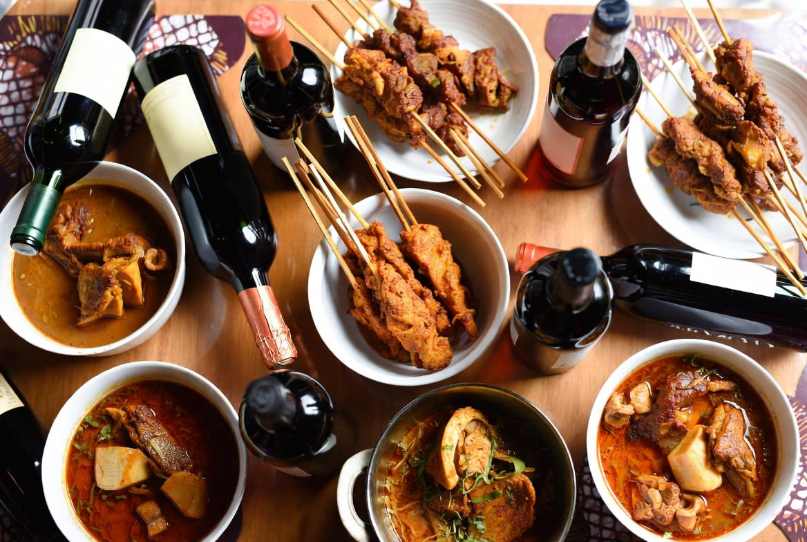 Flat-lay of assorted red wine bottles alongside bowls of egusi soup, suya skewers, and pepper soup on a Nigerian dining table