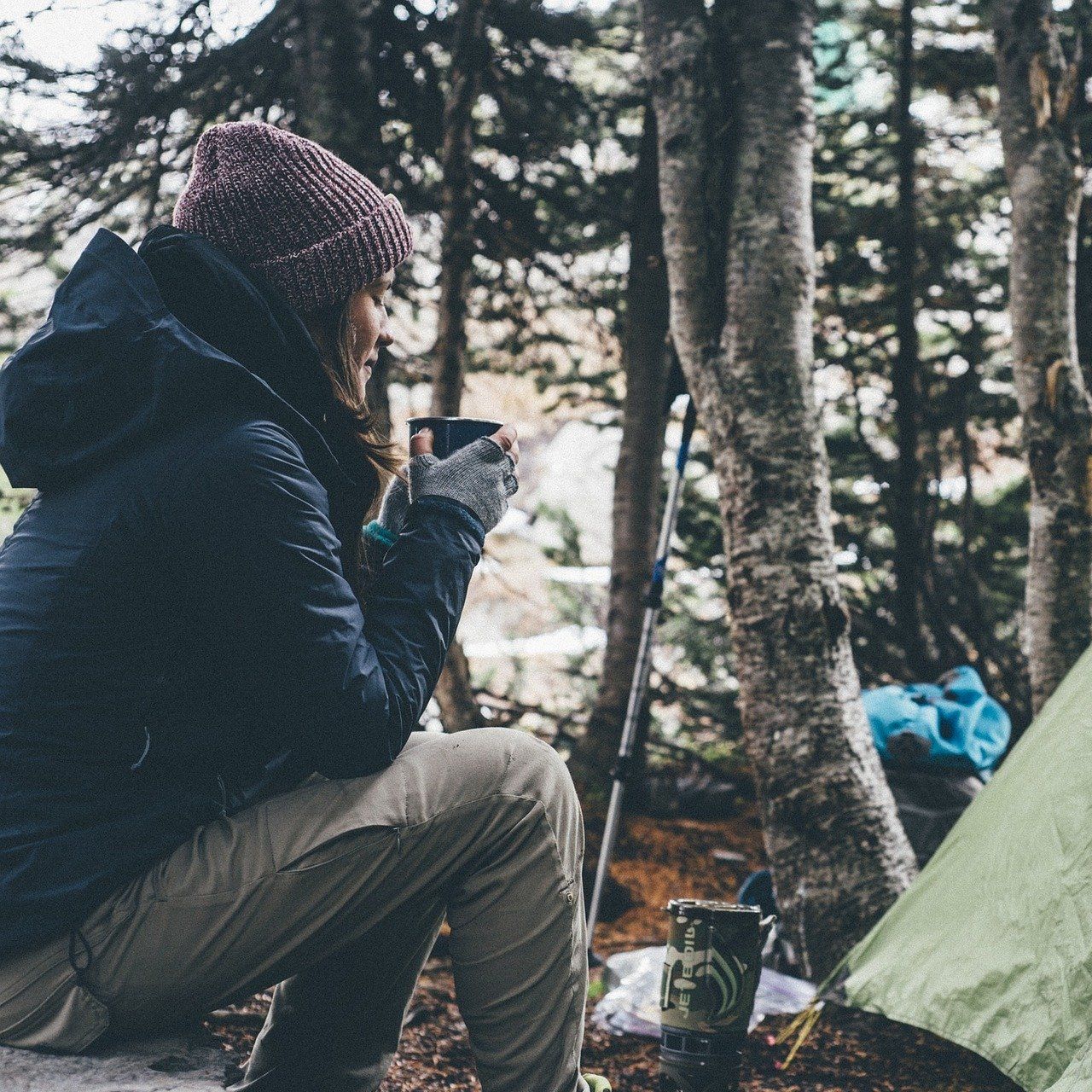 A person sits in front of a tent in a light forest and holds a cup. Trees, camping equipment and a calm natural atmosphere can be seen in the background.