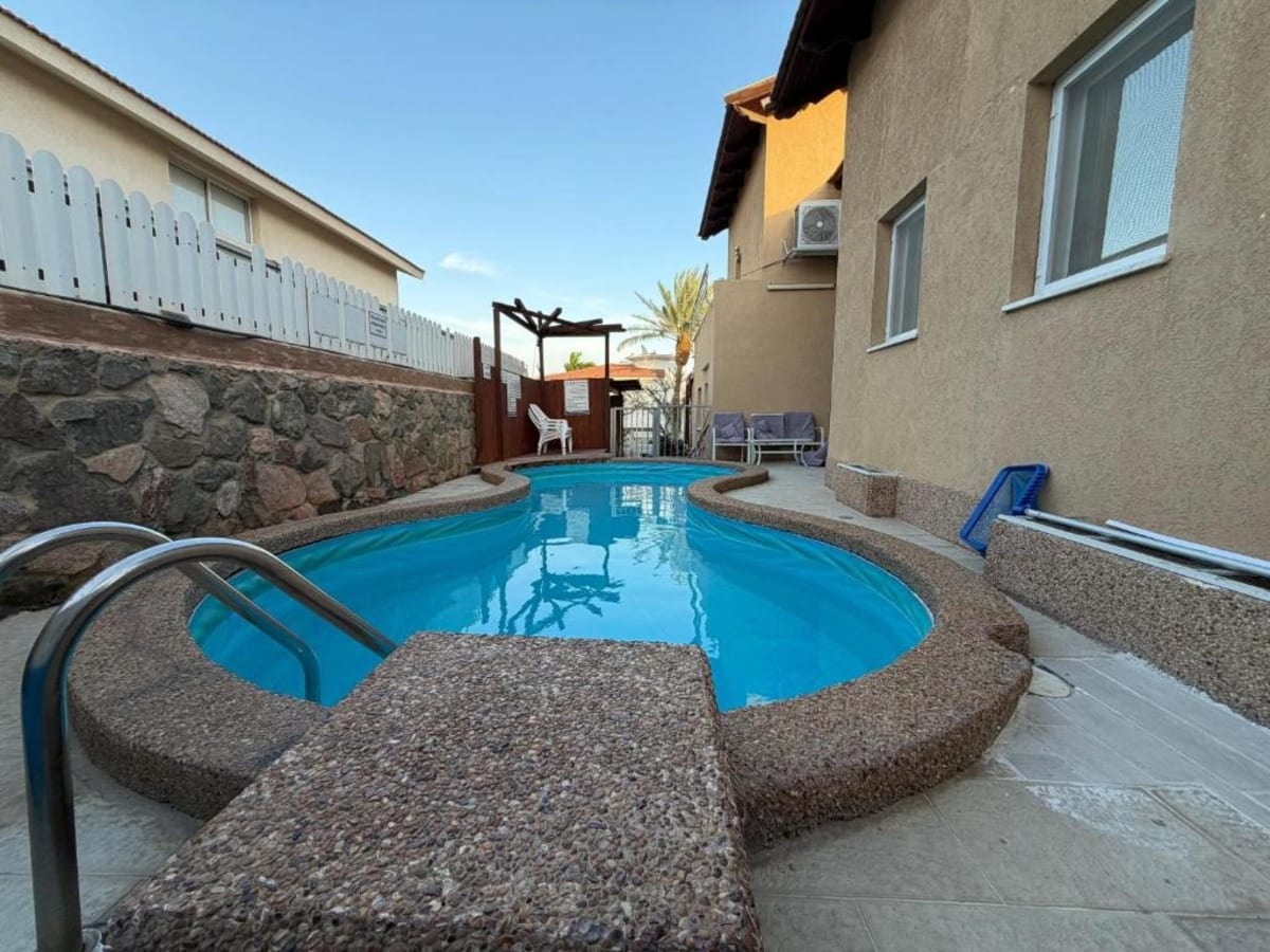 Piscine privée en forme de haricot dans une cour pavée entre les murs de la maison et une clôture en bois blanc, dans une villa à Eilat