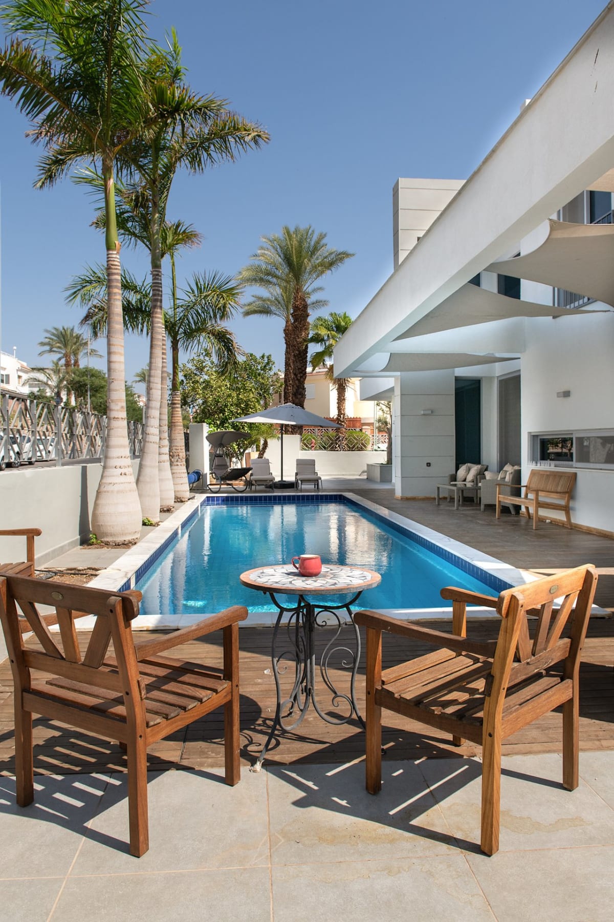 A private pool surrounded by palm trees and a wooden deck, with wooden chairs and a small table, in the yard of a white villa in the Shachamon neighborhood in Eilat