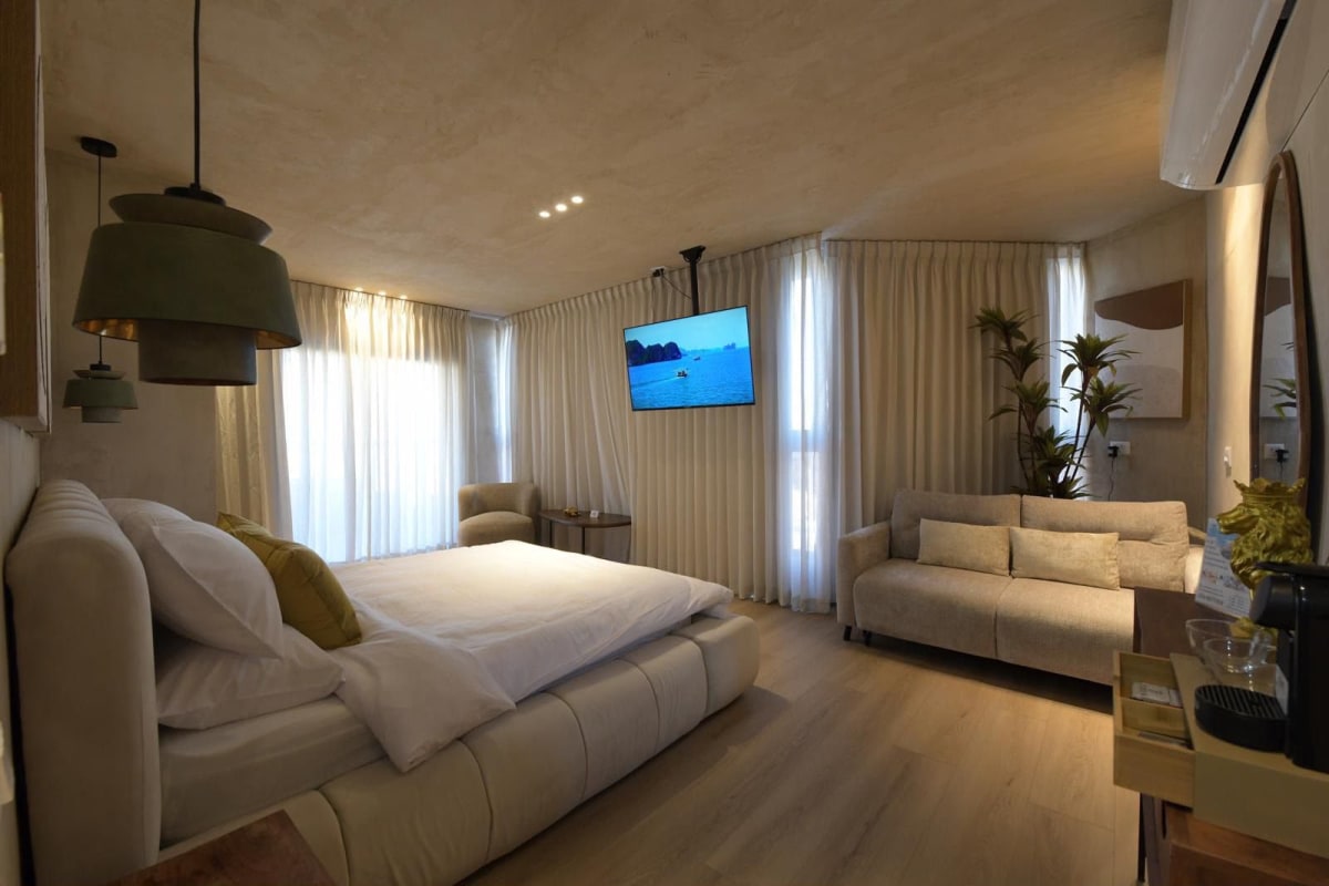 Bedroom in a suite in Eilat city center: double bed, light sofa, wall-mounted TV, and white curtains along the windows