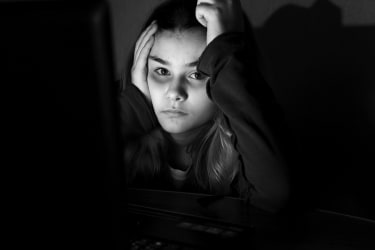 En teenagepige sidder i mørket med hovedet i hænderne og bedrøvede øjne