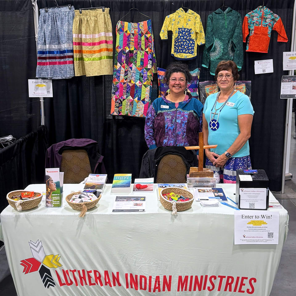 Two Lutheran Indian Ministries staff smiling behind the LIM booth table in the AFN exhibitor hall, with the prize kuspuks, ribbon skirts and patchwork quilt hanging behind them
