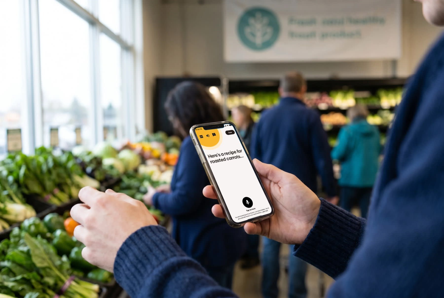 A person using the Nuna app on their phone for a roasted-carrots recipe while shopping in a grocery produce aisle