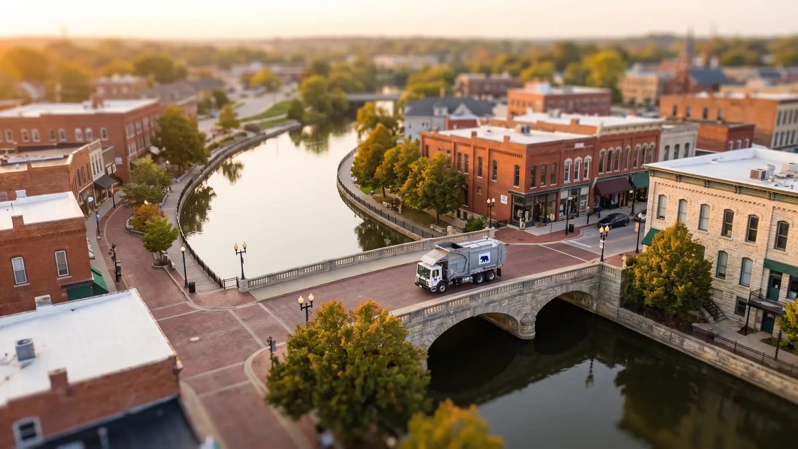 Aerial tilt-shift view of a Fox Valley river town with a Midwest Waste truck crossing the bridge