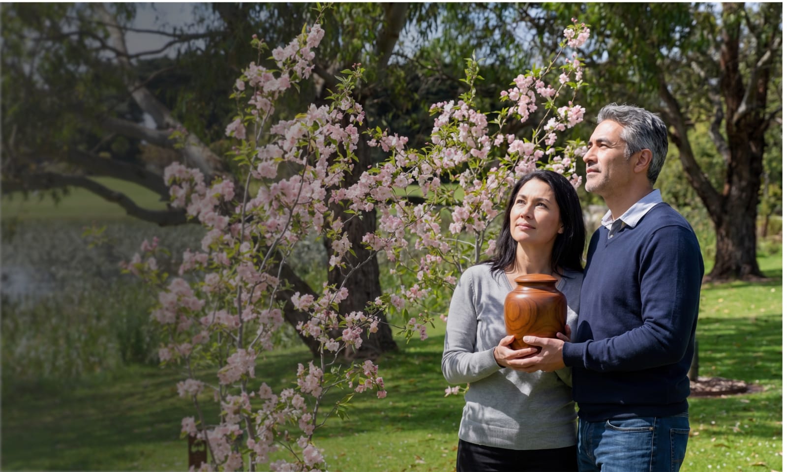 A couple holding a loved one's ashes in a wooden urn among flowering memorial trees at Mornington Green.
