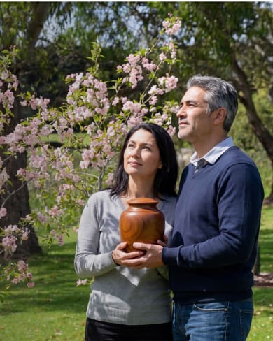A couple holding a loved one's ashes in a wooden urn among flowering memorial trees at Mornington Green.