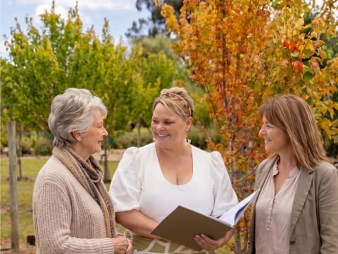 A Mornington Green legacy planner sharing a brochure with two women among the autumn memorial trees.