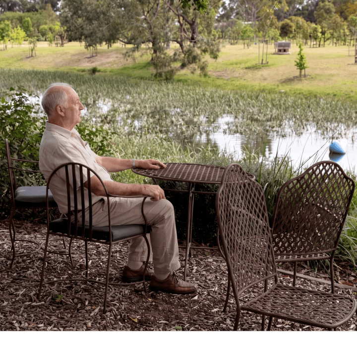 An older man sitting at a table by the lake among the memorial trees at Mornington Green.