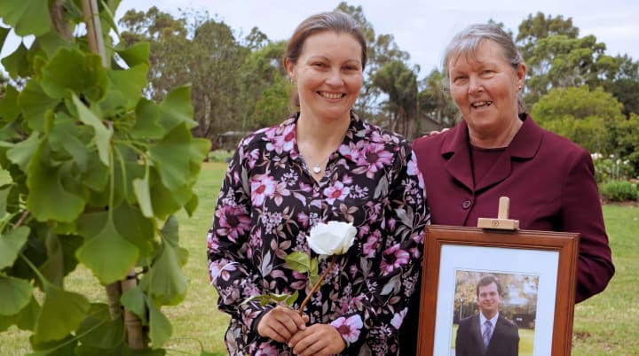 Two women smiling beside a ginkgo memorial tree, one holding a white rose and the other a framed photo of their loved one.