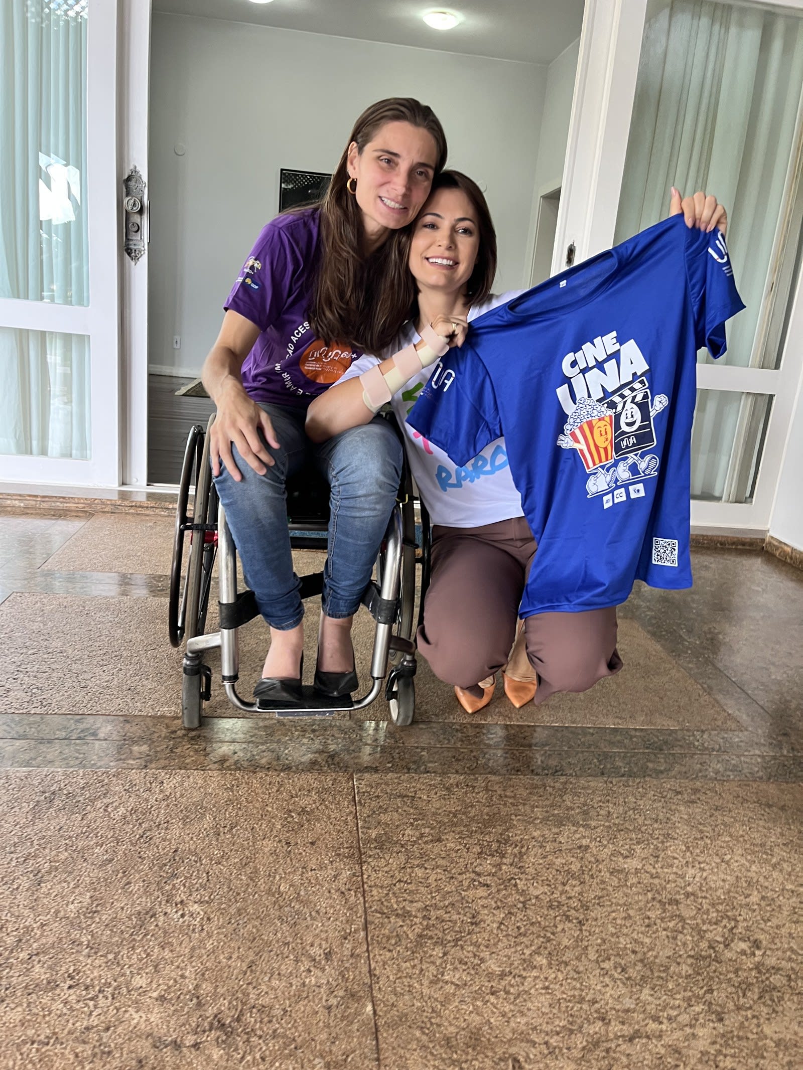 Andréa Pontes, em cadeira de rodas e camiseta roxa, ao lado de Michelle, que segura uma camiseta azul do evento cultural Cine Una