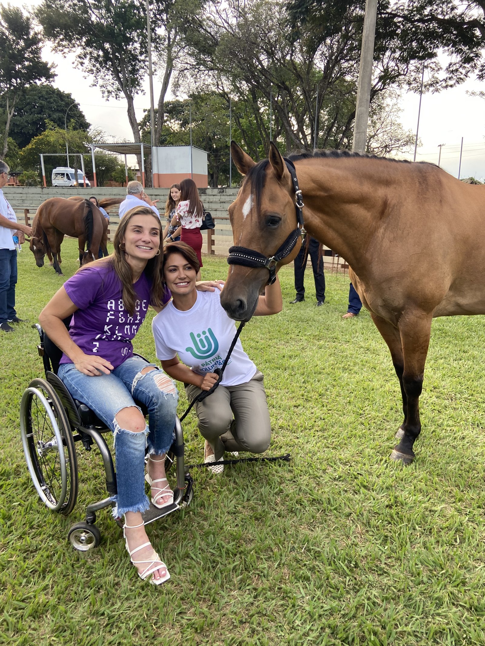 Andréa Pontes, em cadeira de rodas, ao lado de uma voluntária da Una e de um cavalo durante uma sessão de equoterapia ao ar livre