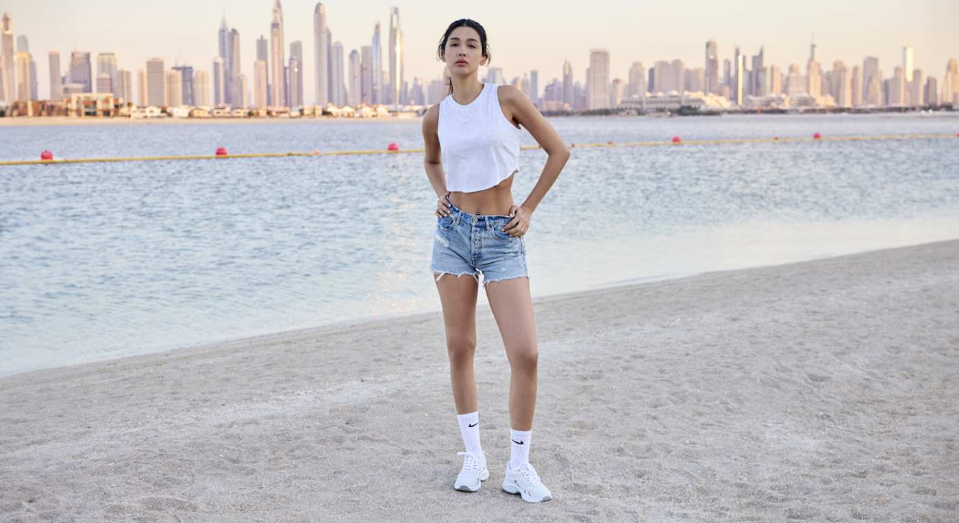 Frau steht in Athleisure-Style-Outfit am Strand, Skyline von Dubai im Hintergrund