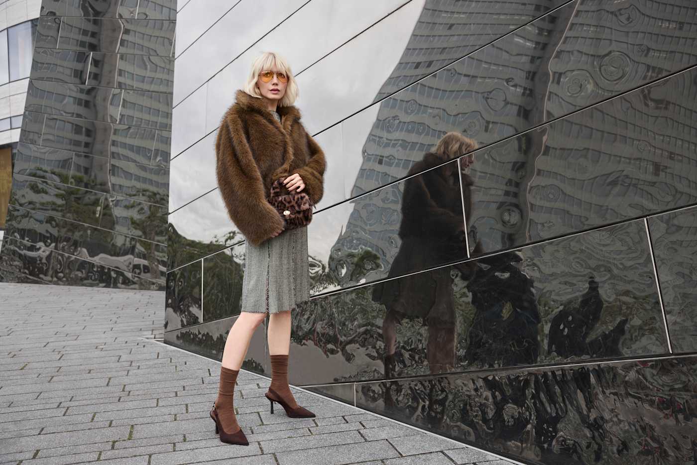 Frau trägt braune Felljacke im Boho-Style, mit Strickkleid und Slingback-Pumps vor Spiegelfassade