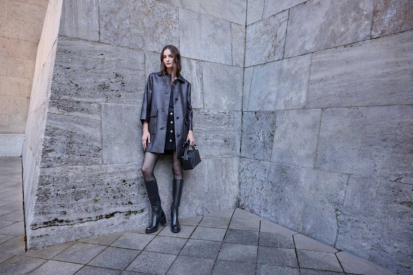 Frau in grauem Mantel und Kleid vor Steinwand kombiniert schwarze Kniestiefel mit Blockabsatz und Minitasche