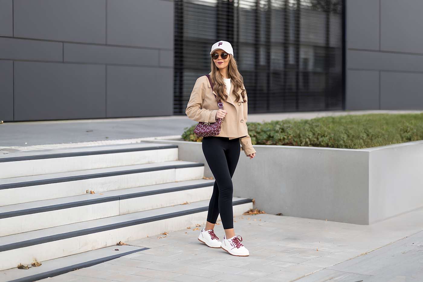Frau in braunem Cropped Trenchcoat und schwarzer Leggings, mit Handtasche und FILA-Cap