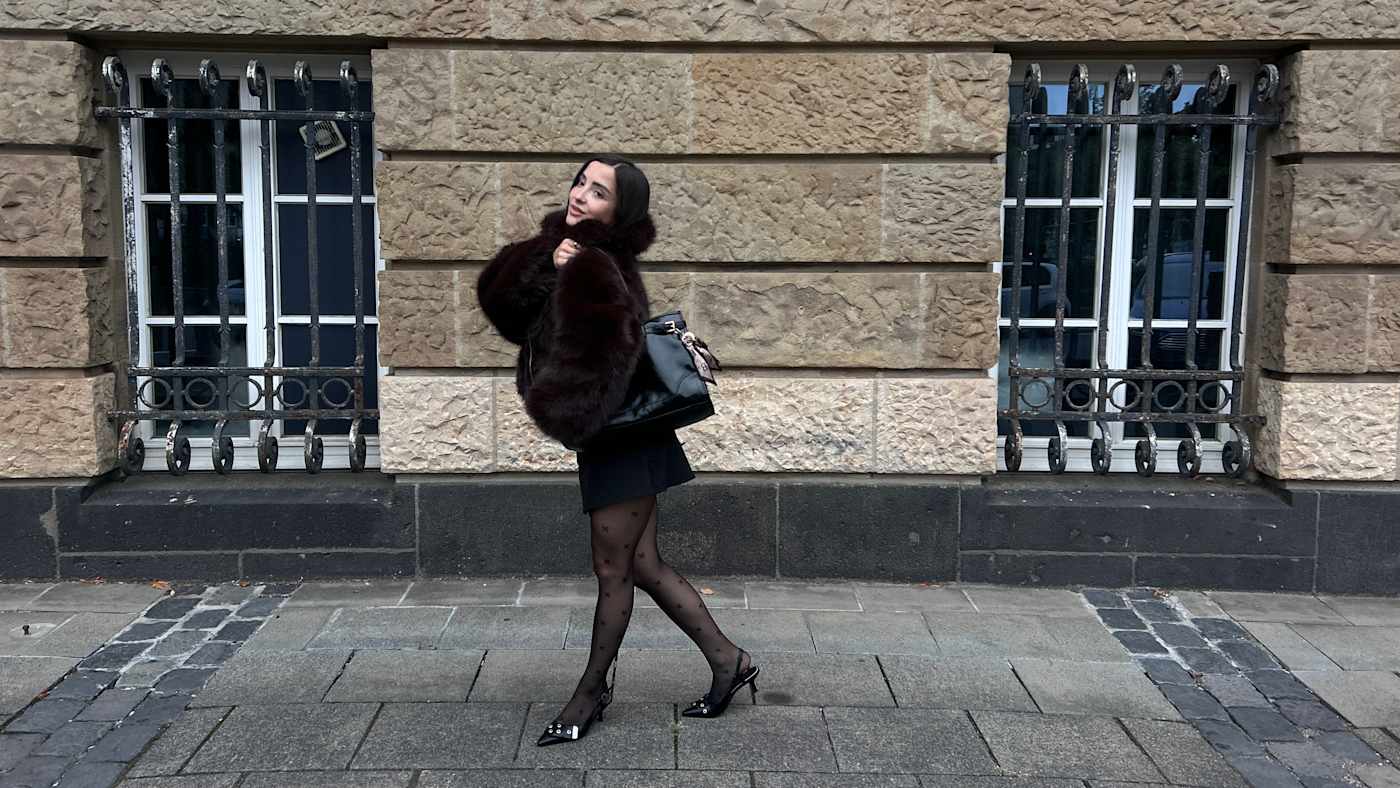 Frau in dunklem Pelzmantel mit schwarzer Handtasche und Strumpfhose, steht Weg vor Gebäude