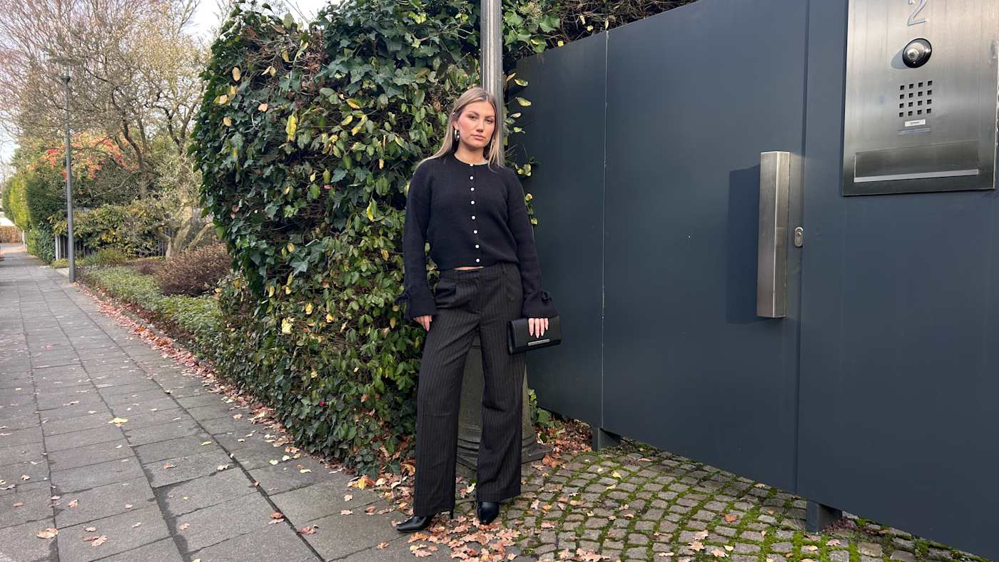 Junge Frau in schwarzem Cardigan und schwarzer Hose, steht vor Gartentor