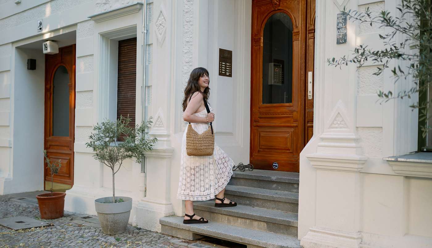 Frau in weißem Hemdkleid, trägt beige Basttasche, steigt Treppenstufen