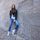 Model in Lederjacke und Perlen-Jeans mit weißen New-Balance-Sneakern vor Steinwand